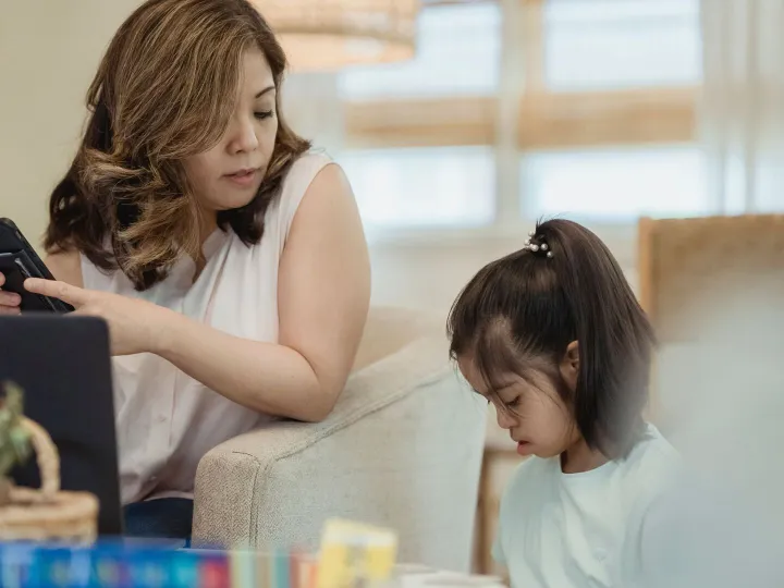 Parent looking at her phone at home while her young daughter plays beside her