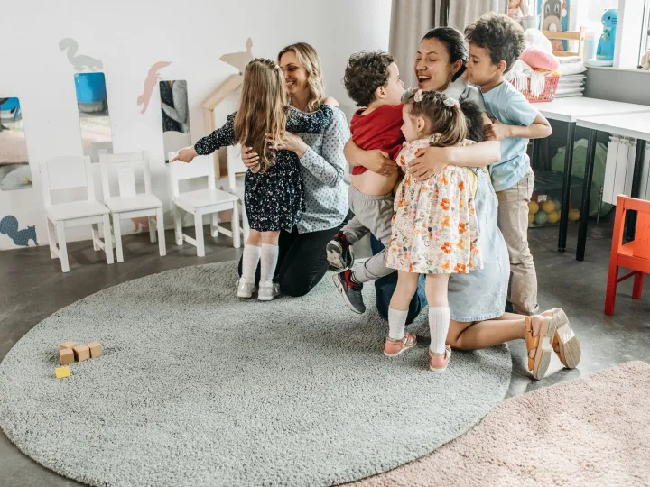 Young children hugging two nursery practitioners in a playroom