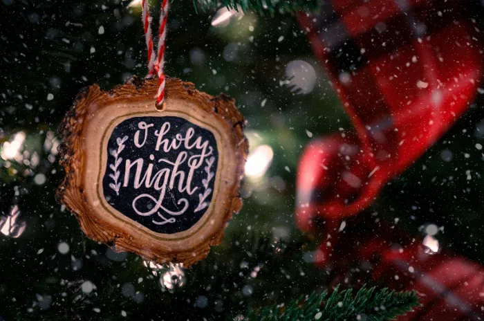 Wooden bauble with O Holy Night inscription hanging on green Christmas tree decorated with red ribbon on snowy day