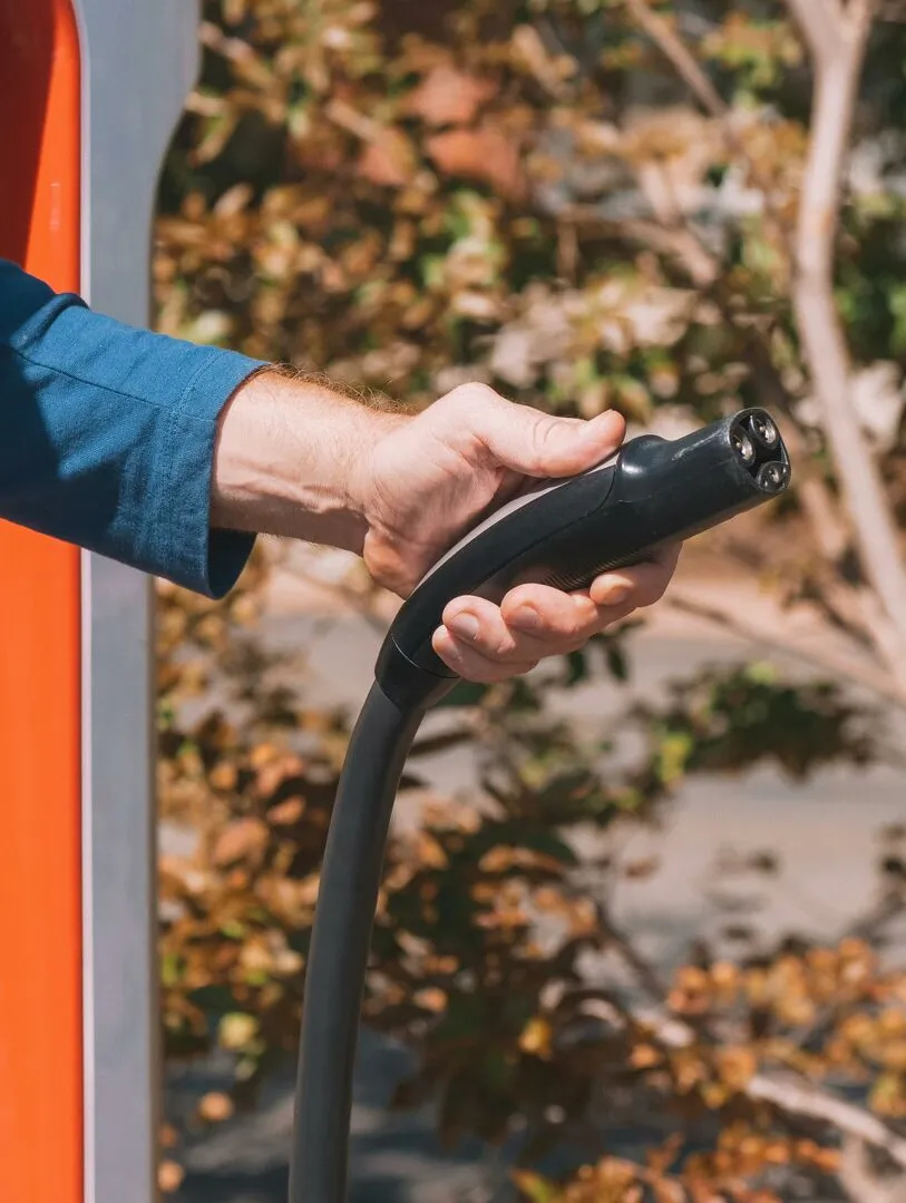 Hand holding an electric vehicle charger at a station outdoors with autumn trees.