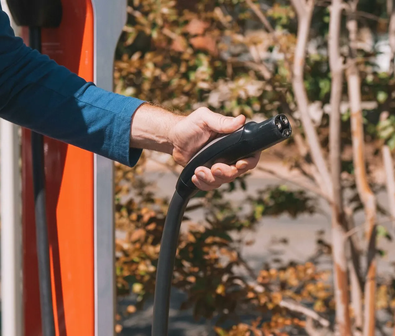 Hand holding an electric vehicle charger at a station outdoors with autumn trees.