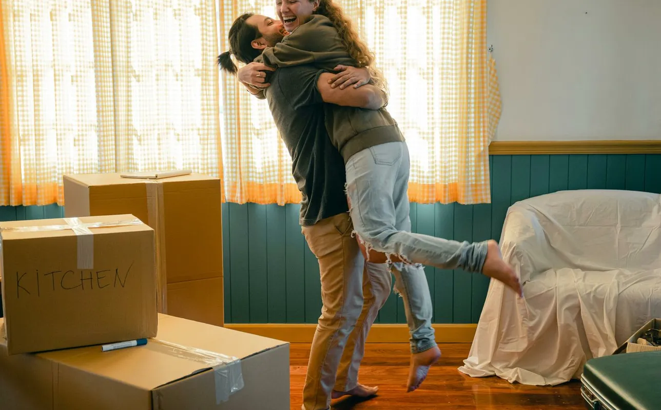 Side view full body barefoot young bearded male in casual clothes standing against window and lifting laughing girlfriend up during relocation and unpacking things from cardboard boxes