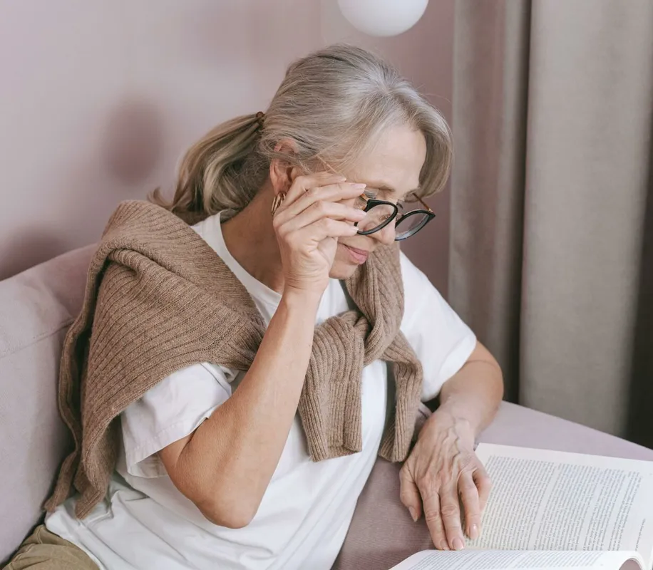 Elderly woman with glasses reading on a couch for leisure at home.