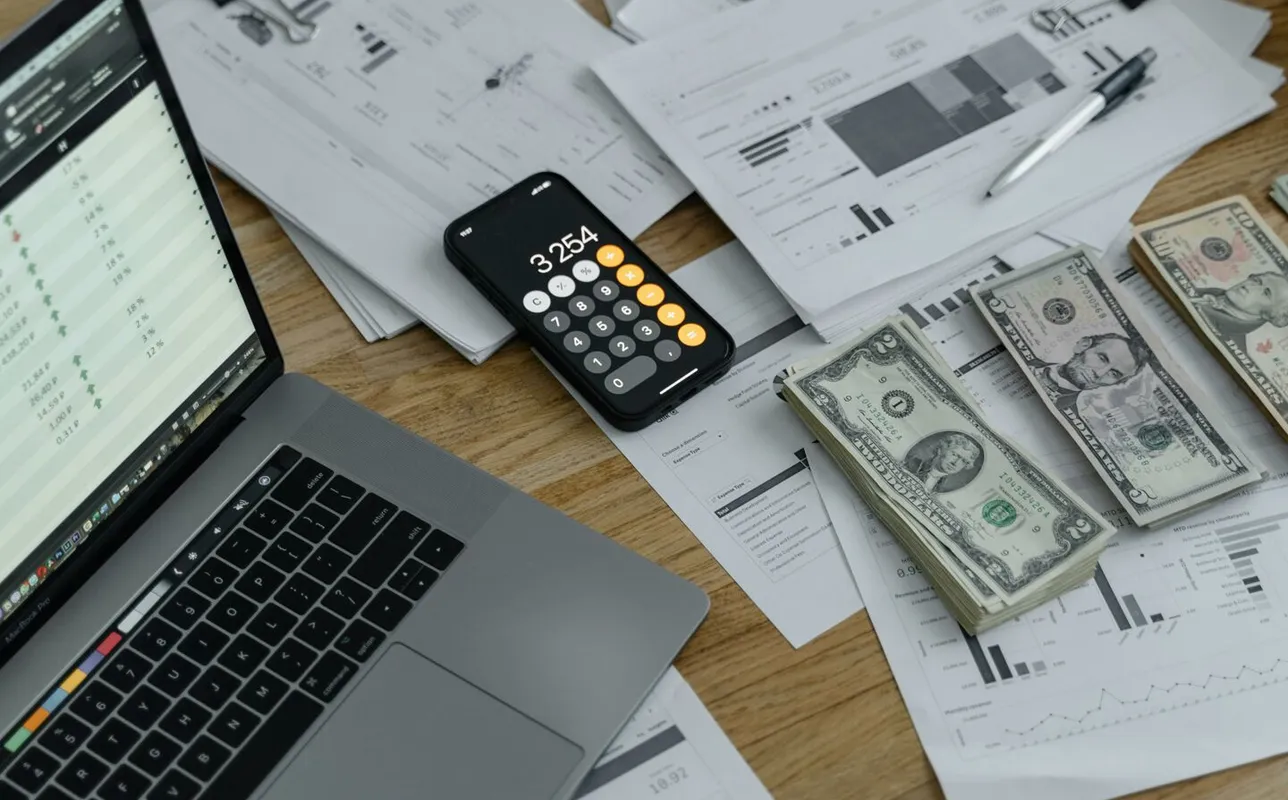 A desk with financial documents, currency, a laptop, and phone calculator.