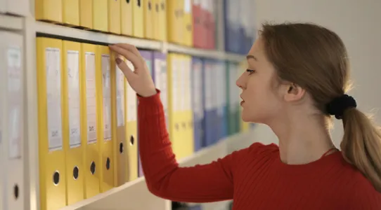 Side view of contemplating female assistant in casual style standing near shelves and choosing file with documents