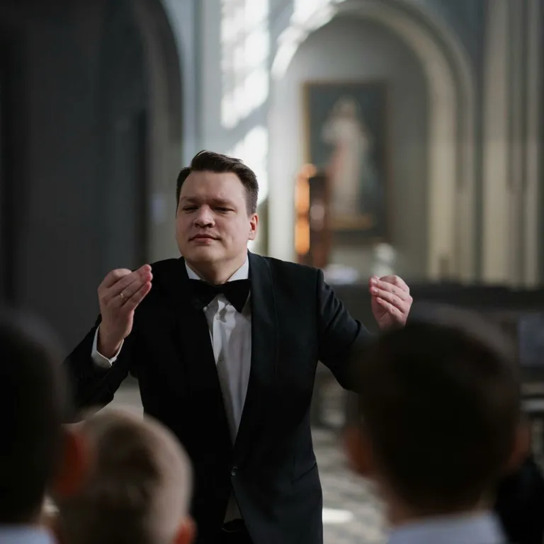 A male conductor leads a boys' choir rehearsal in a serene church setting.