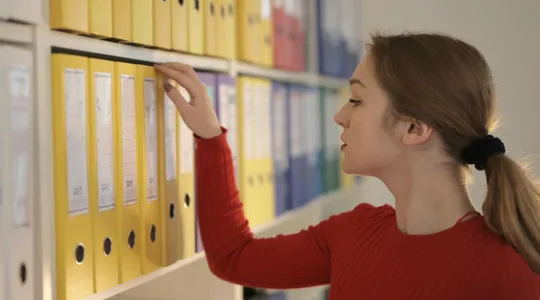 Side view of contemplating female assistant in casual style standing near shelves and choosing file with documents