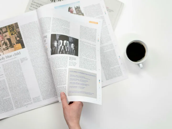 A close-up shot of a hand holding a newspaper with a coffee cup on a white surface.