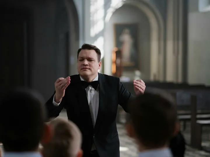 A male conductor leads a boys' choir rehearsal in a serene church setting.