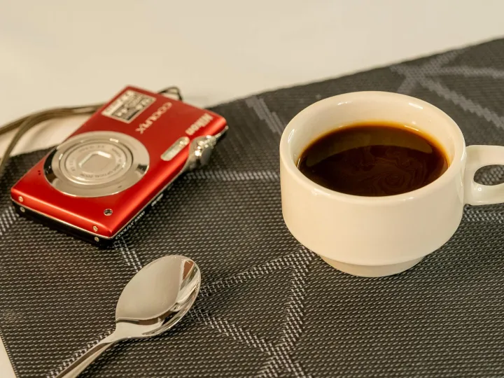 A white cup of coffee next to a red camera and spo