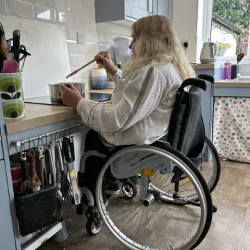 A woman sitting in a wheelchair is stirring a saucepan. The hob is height adjustable, with space below to allow her to sit close. The kitchen units are pale blue with wooden worktops. She has long blonde hair and is wearing a white top.