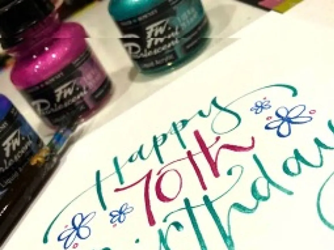Happy 70th birthday card lettered in blue and pink beside pots of ink