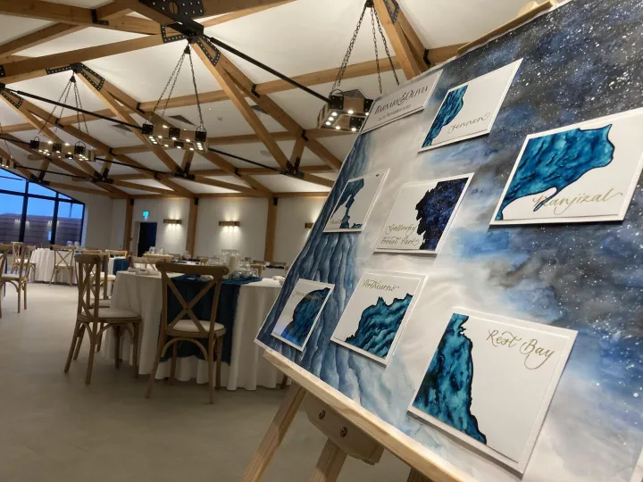 Hand lettered wedding table plan with a galaxy watercolour background and coastal place names, displayed on an easel in a timber framed barn