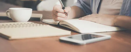 Woman Writing on a Notebook Beside Teacup and Tablet Computer