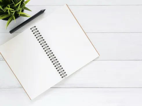 Open spiral notebook on white desk with black pen and green plant. Ideal for planning.