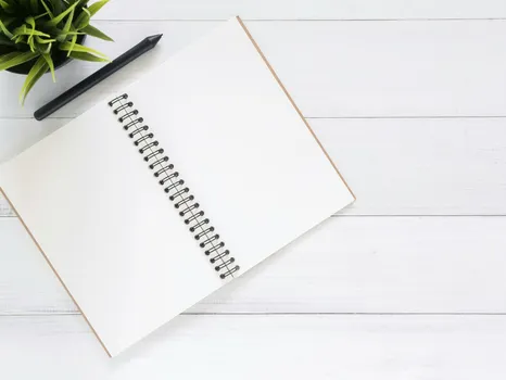 Open spiral notebook on white desk with black pen and green plant. Ideal for planning.