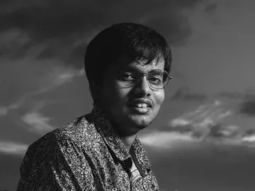 A black and white portrait of a smiling man with eyeglasses and dark hair, set against a dramatic sky.