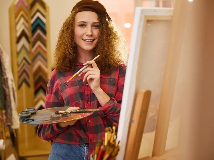 Young curly artist looks happy and holds a palette with oil 