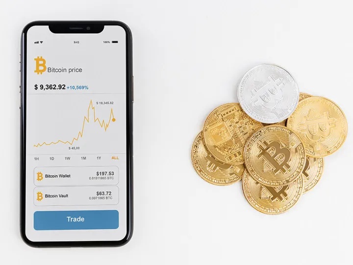 Smartphone displaying live bitcoin price next to gold and silver cryptocurrency coins on a white background